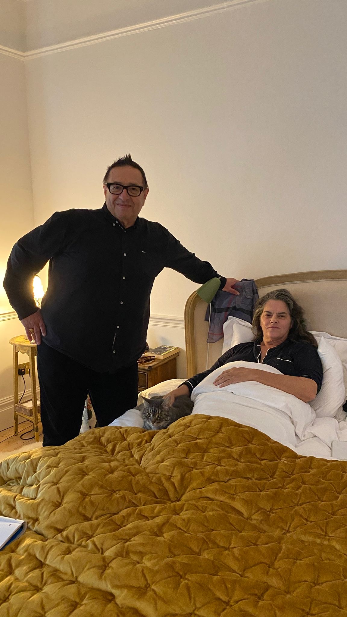 A man standing next to a woman and cat in bed.