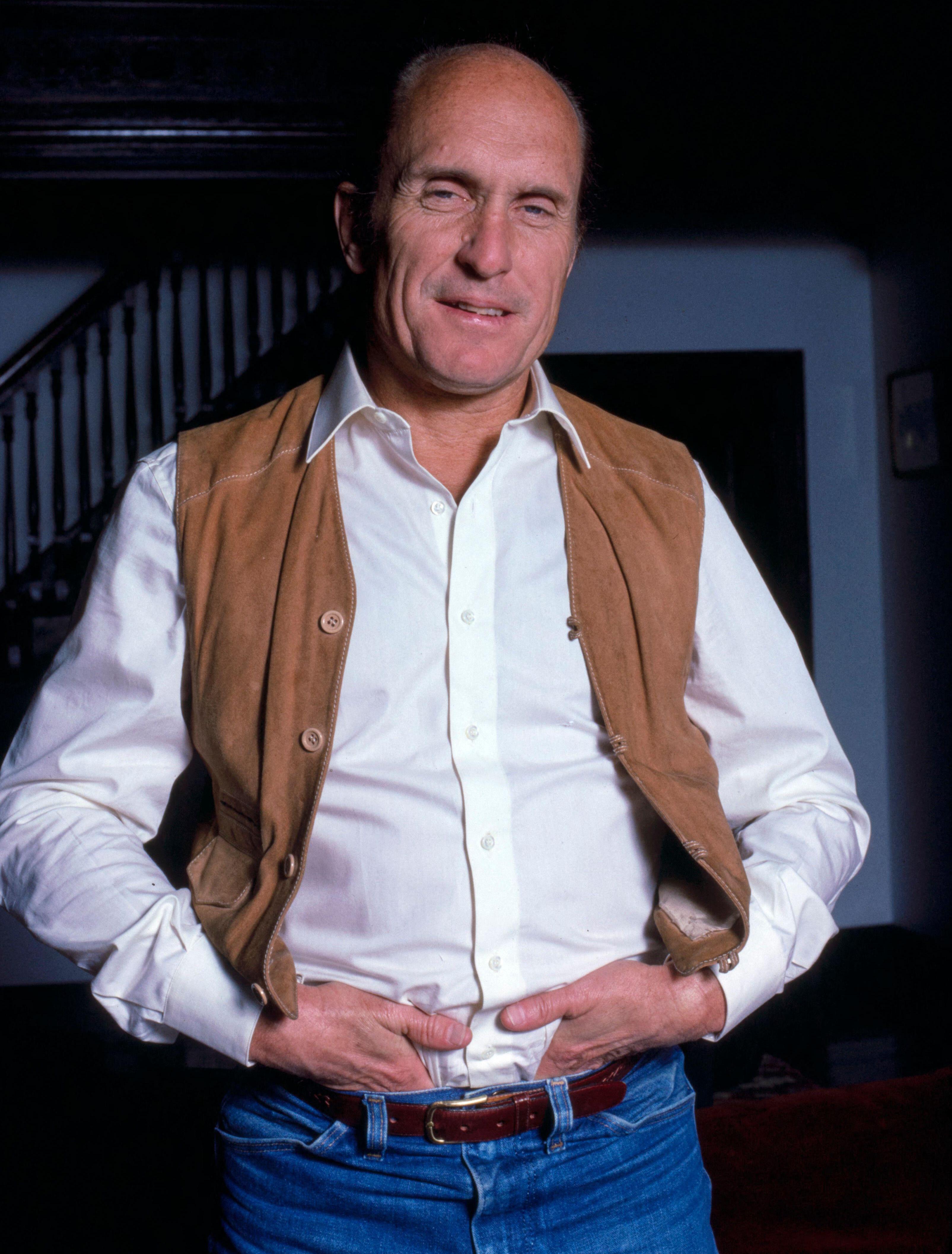 Robert Duvall, an American actor and filmmaker, stands smiling in a white shirt, tan vest, and jeans at his New York City home.