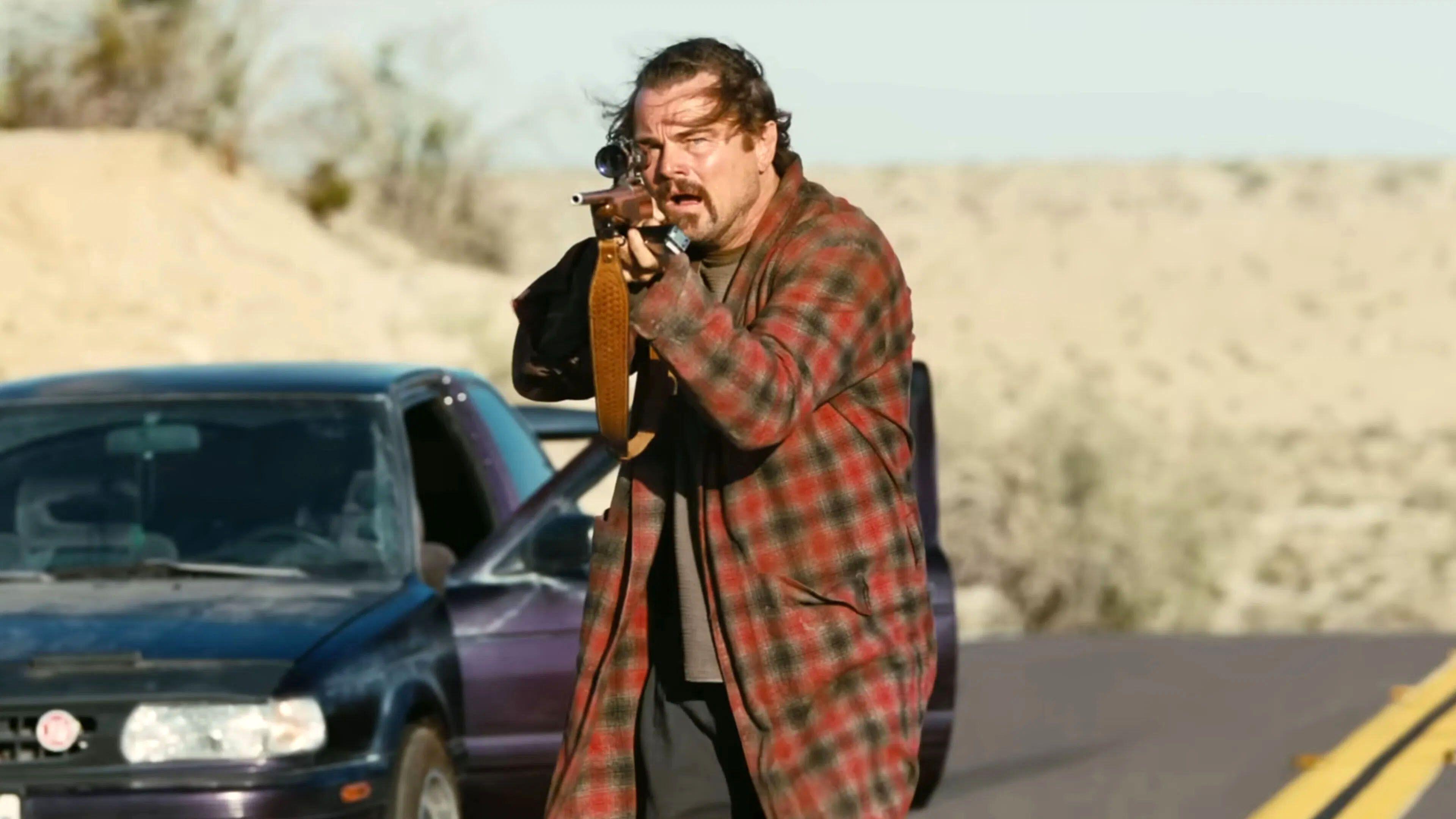 Leonardo DiCaprio holding a rifle, dressed in a plaid coat, with a car in the background, in "One Battle After Another."
