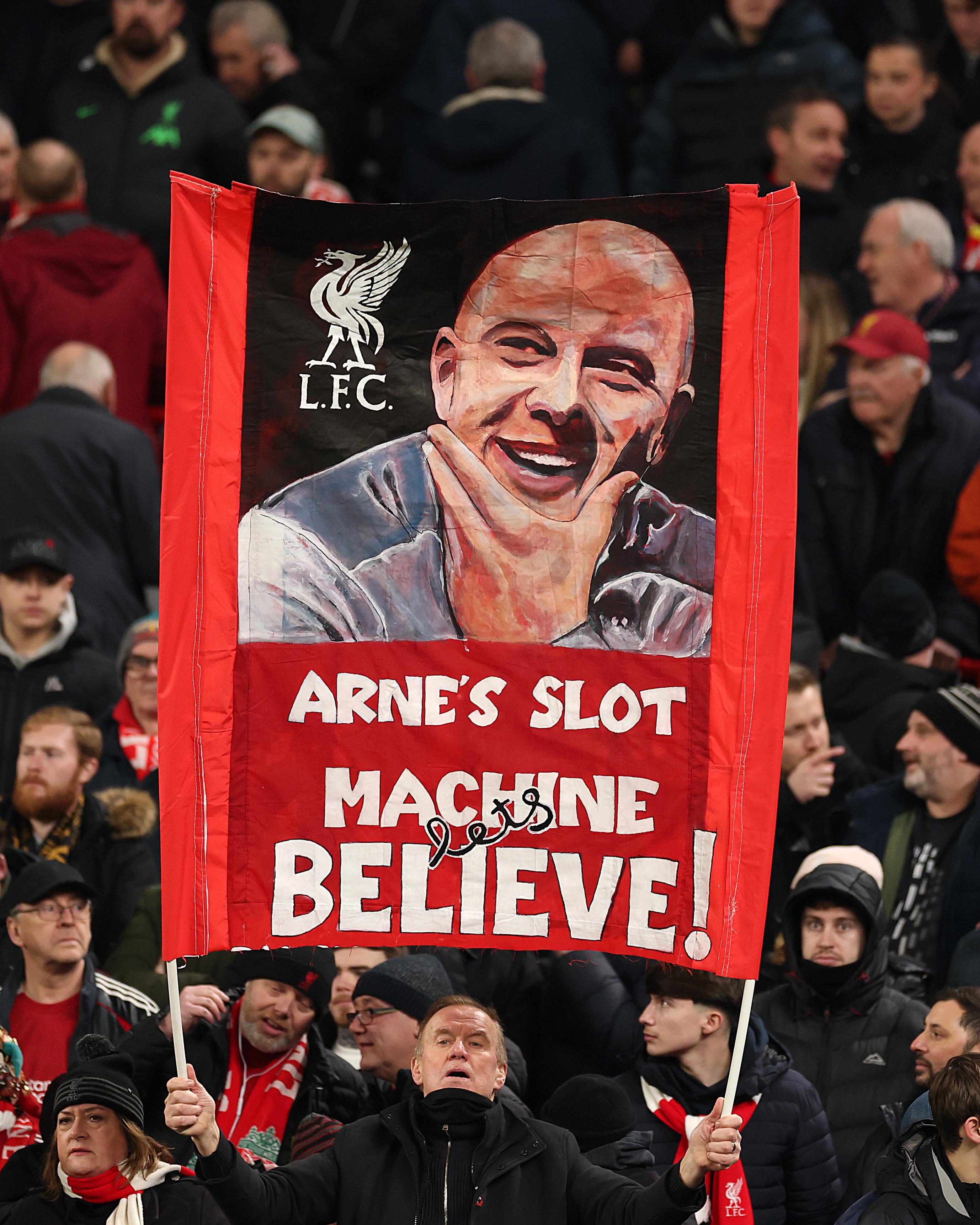 A fan holding a flag with a caricature of Arne Slot and text "Arne's Slot Machine Let's Believe!"