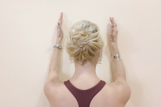 A woman with blonde hair in a bun stands facing away, with her arms raised and palms pressed against a wall.