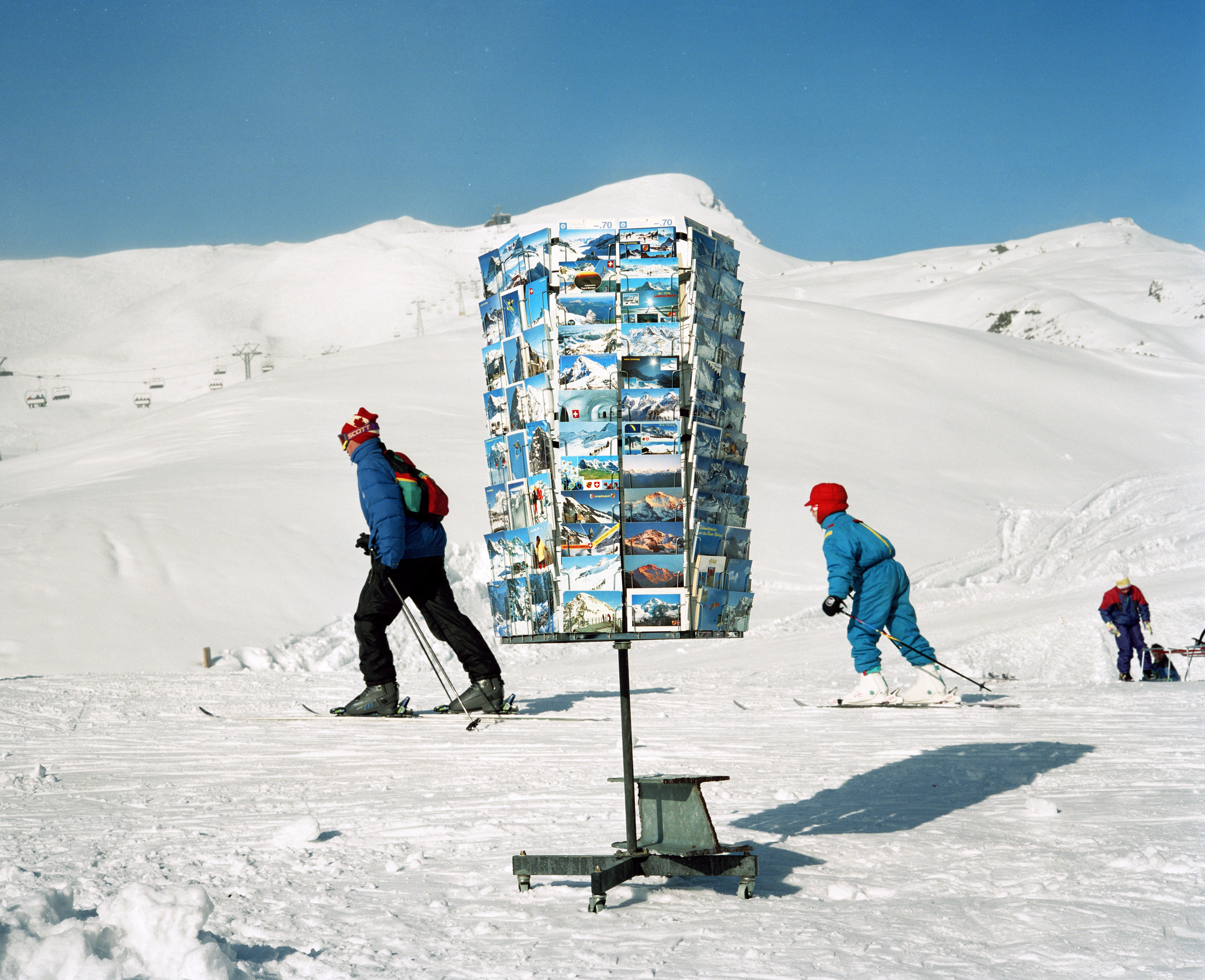 A postcard display stand on a snow-covered mountain with skiers in the background.