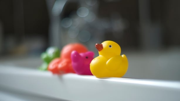4 bright coloured rubber bath toys yellow,pink, orange and green sitting on side of bath, yellow rubber duck in foreground
