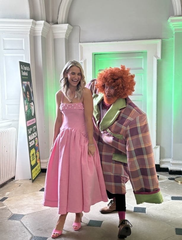 A blonde woman in a pink dress laughs beside a man dressed in oversized clothes and wearing a red curly wig