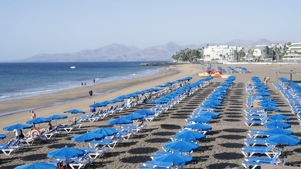 Wide angle view of tourists sunbathing on sun loungers under rows of blue umbrellas on a long sandy beach in Puerto del Carmen in Lanzarote in the Canary Islands in Spain on November 22, 2025. The scene shows holidaymakers walking along the shoreline with hotels and palm trees in the background and 