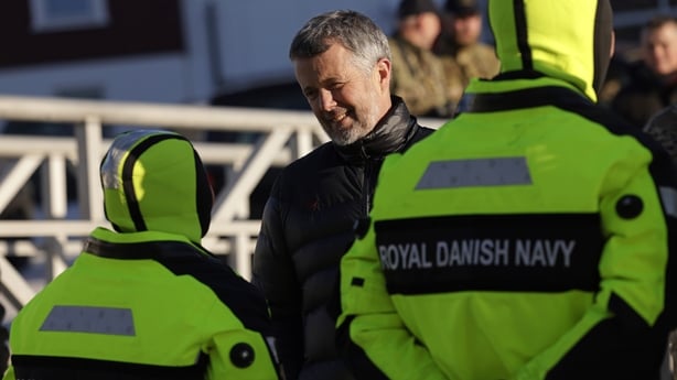 a smiling man speaks to two people wearing uniforms with 'royal danish navy' written on them