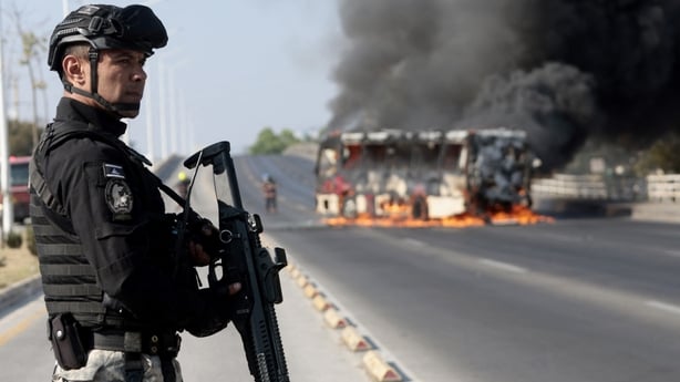A member of the Prosecutor's Office stands guard near a burning bus at one of the main avenues after it was set on fire by organised crime groups in response to an operation in Jalisco to arrest a high-priority security target in Zapopan, state of Jalisco, Mexico, on February 22, 2026. Armed civilia