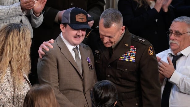 a man in uniform puts his arm around another man wearing a baseball cap as other people look on and clap