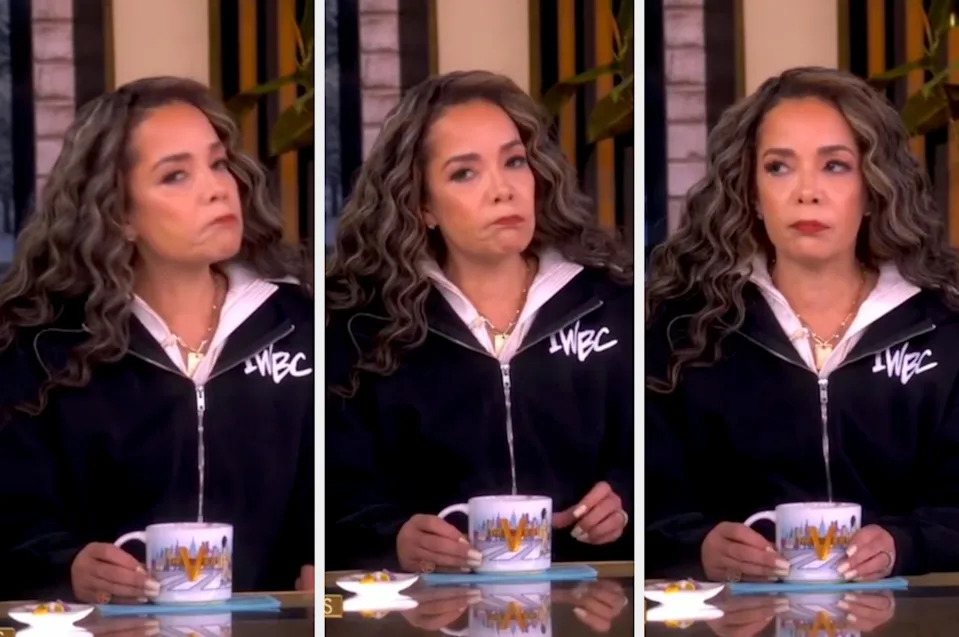 A person on a talk show holds a mug, wearing a casual zip-up top with curly hair. The expression changes slightly, showing contemplation or concern