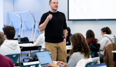 Matthias Schlichting, wearing a black sweater and tan pants, stands among his students, leading class.
