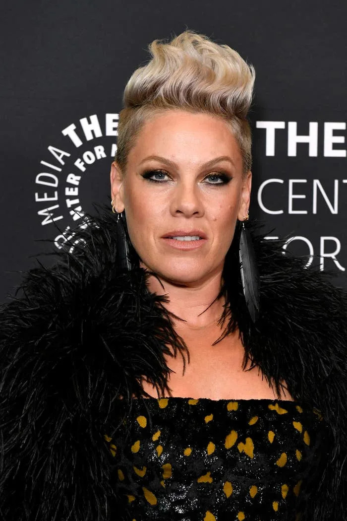 Pink in a feathered jacket and patterned dress at a media event, with a bold hairstyle and dramatic earrings