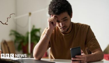 A young man looks at his phone while sitting at a computer in his home. He looks weary.