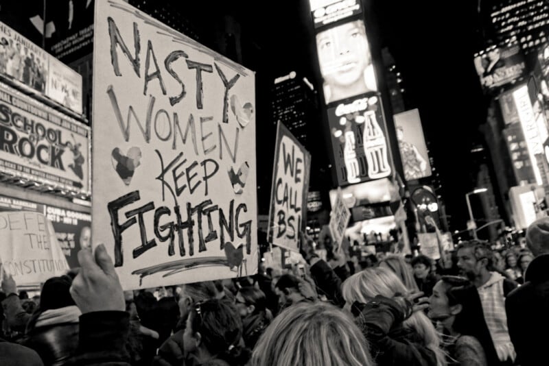 A crowd of protesters at night holds signs, including one that reads “Nasty women keep fighting,” in a brightly lit city area filled with billboards. The atmosphere is energetic and determined.