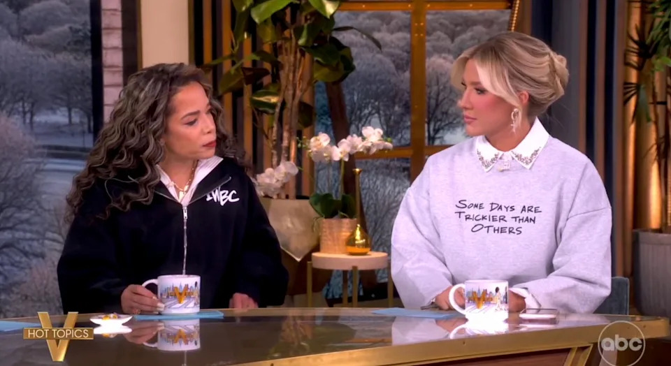 Two people sit at a talk show desk holding mugs, wearing sweatshirts with embroidered designs