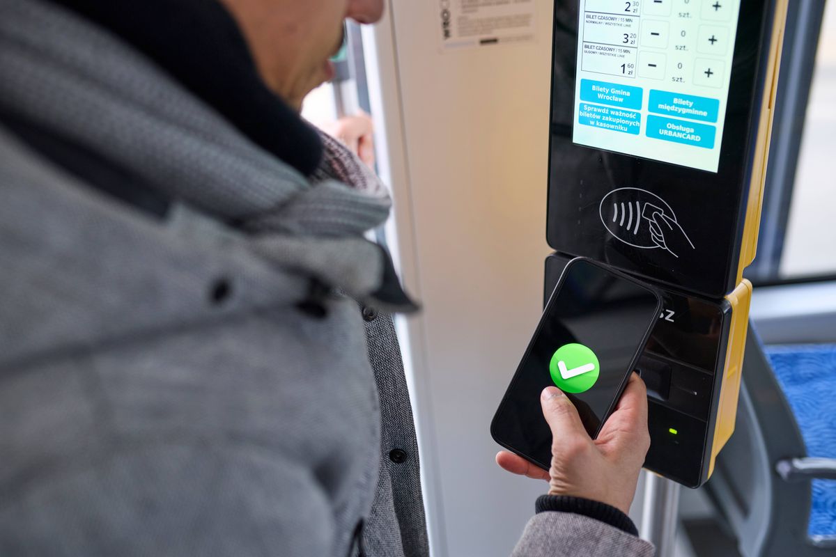 Close-up showing a man in a gray winter jacket holding his smartphone with a green checkmark screen against a yellow contactless ticket reader inside a tram, with blue seats and soft daylight in the background. The scene highlights seamless mobile payment and digital ticketing in urban public transport. Concepts: mobile ticket validation, contactless transit payment, digital commuting convenience