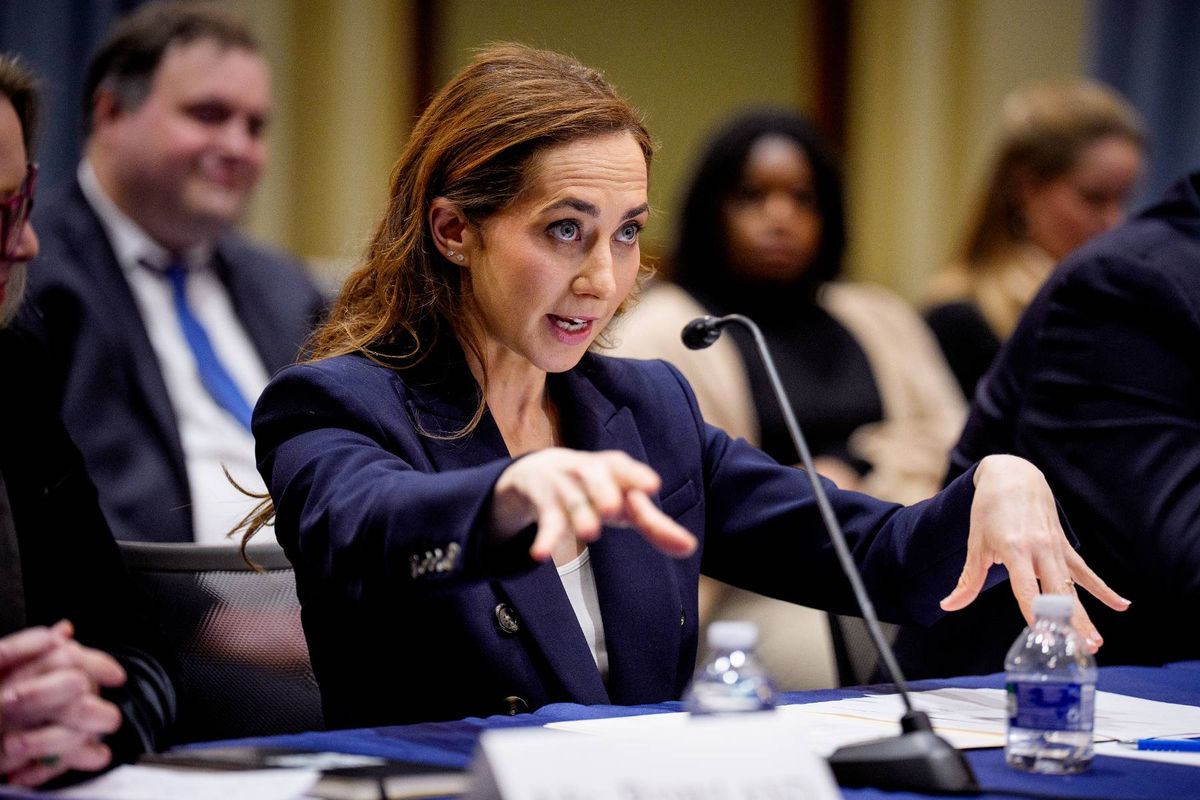 A formal meeting is underway, with an individual seated at a table, actively gesturing and addressing the audience through a microphone. The setting includes a table with various items, and several attendees are seated in the background, attentively listening.
