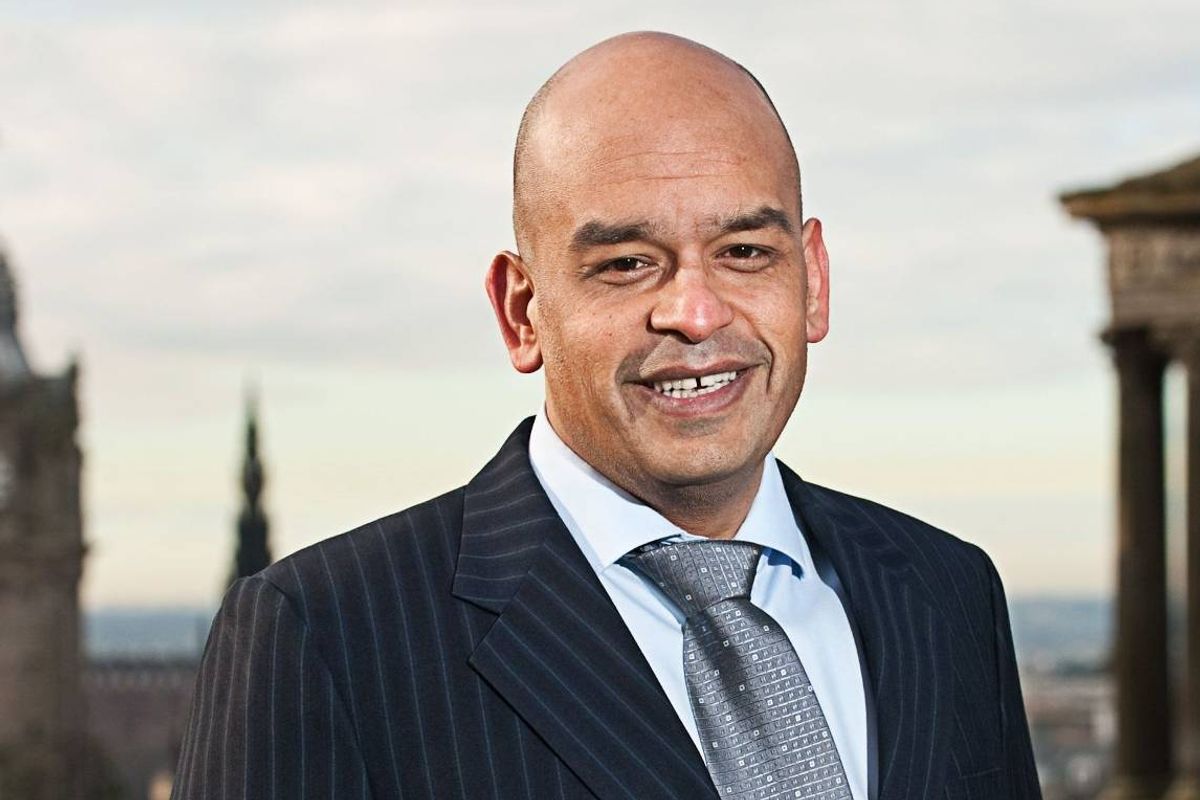 An individual dressed in formal attire, comprising a pinstripe suit and a gray tie, stands confidently with a smile against the backdrop of a city skyline featuring historical architectural elements.