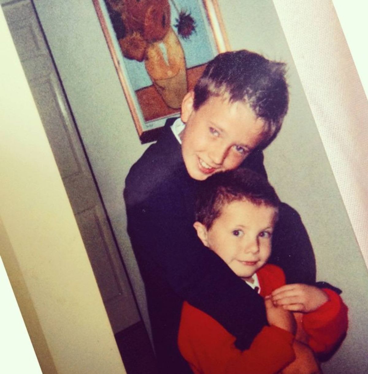 Two young individuals, one slightly taller and older, stand closely together, with the taller one embracing the younger one in a room. Both appear to be smiling, suggesting a moment of camaraderie or affection. The setting is an indoor space with a white wall in the background.