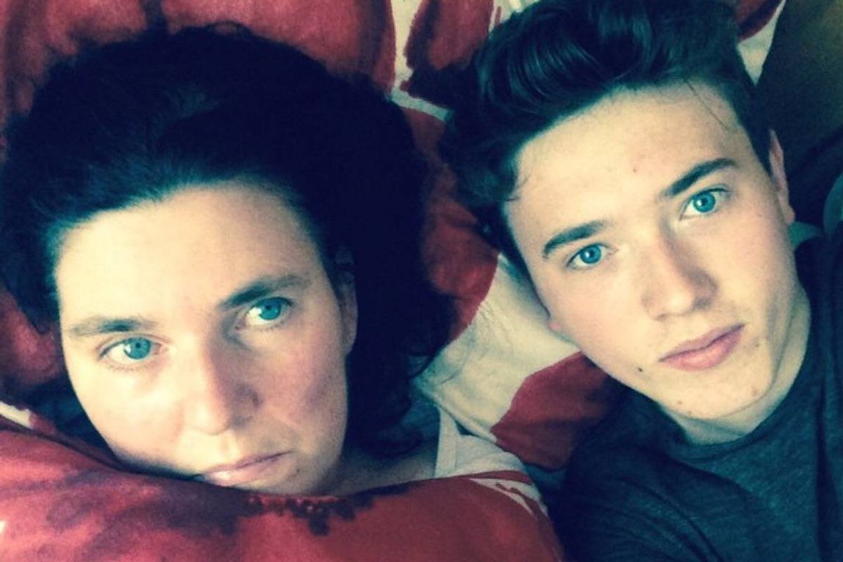 Two individuals, one female and one male, are lying together on a bed with pillows, smiling at the camera. The female has dark hair and green eyes, while the male has short dark hair and light eyes. They appear to be posing for a photograph.