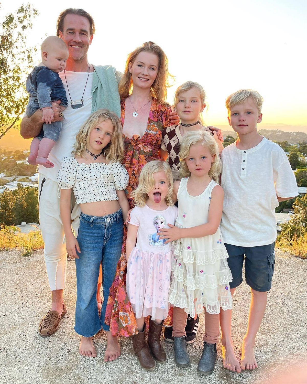 An image depicting a family of seven individuals gathered together for a portrait. They are standing closely, with two adults in the front and five children behind them. The family is dressed in light, casual clothing, and the setting appears to be outdoors with a natural backdrop, possibly a hill or a sunset.