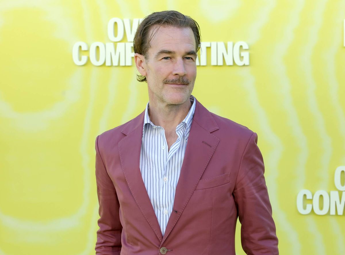 An individual dressed in a burgundy suit and light blue shirt stands against a bright yellow backdrop, likely at a public event or premiere. The background features the text "COMING" and "COM" in white font.