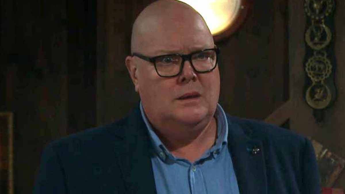 An individual with a bald head, wearing glasses and a formal suit, appears to be engaged in a conversation, possibly in a professional setting.