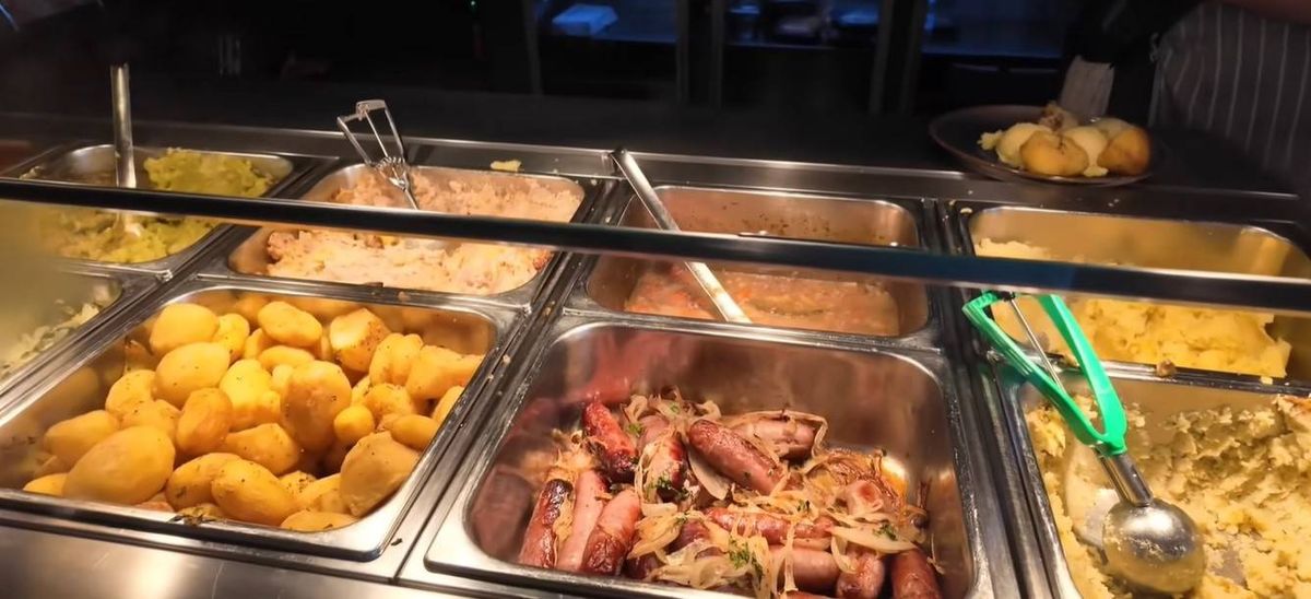 An image displaying a buffet table with various food items in stainless steel containers, including a dish of noodles and meat, accompanied by utensils for serving.