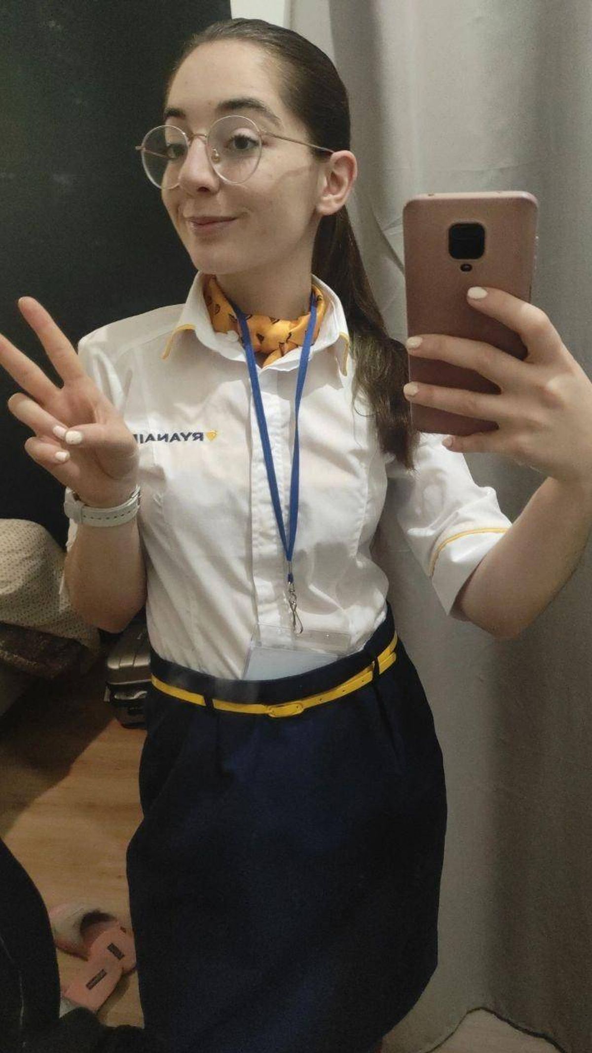An individual in a white shirt with a blue lanyard around their neck is making a peace sign while holding a smartphone, presumably taking a selfie.