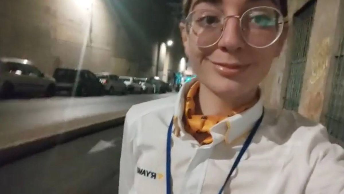 An individual, dressed in a white lab coat with a blue lanyard, stands confidently on a city street at night, wearing glasses and a hint of a smile. The background features an urban setting with parked cars and street lighting.