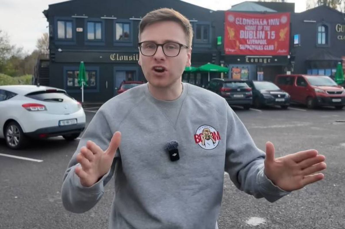 An individual in a gray sweatshirt stands in an outdoor area, gesturing with their hands, with a building and parked cars in the background.