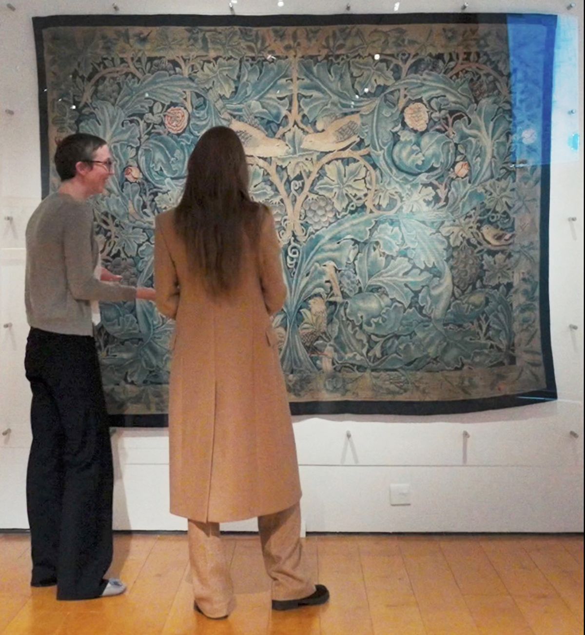 Kate admires a large textile featuring floral and leafy patterns.