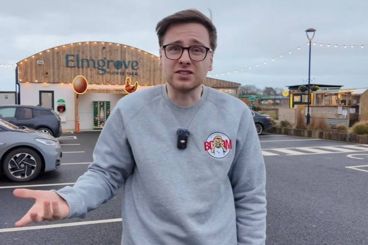 A man wearing a gray sweatshirt and glasses stands in a parking lot, gesturing towards a building under construction. Several vehicles are parked nearby, and streetlights are visible in the background.