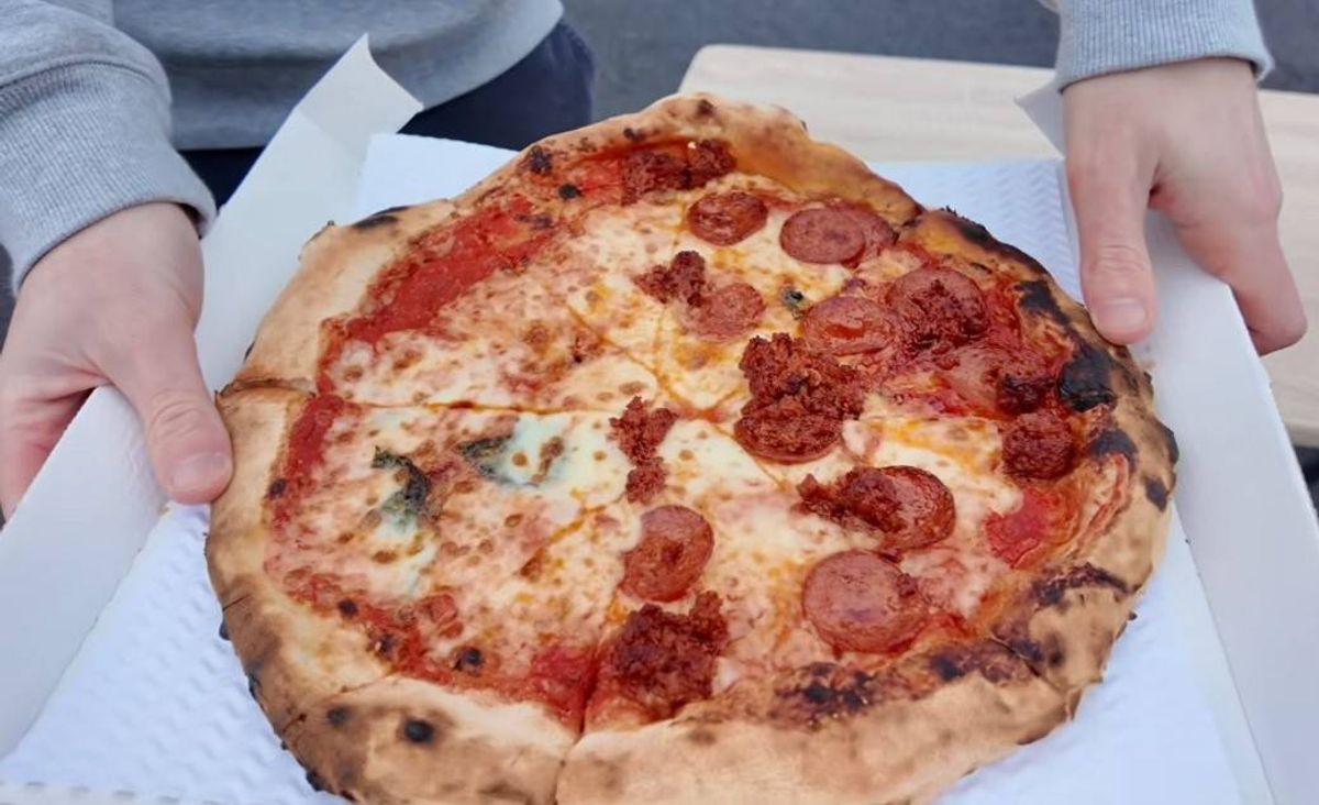 An individual is holding a freshly prepared pizza, complete with melted cheese, pepperoni, and a few fresh herbs on top, positioned within a white paper box.