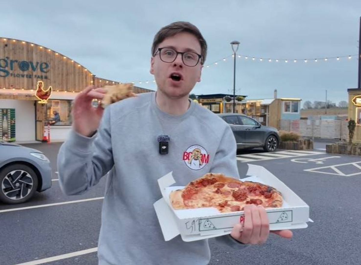 A person holding a pizza box in an open area with a building and vehicles in the background.