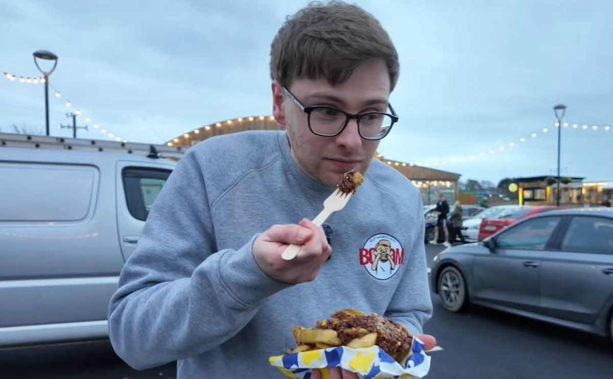 An individual in a grey sweatshirt, donning glasses, holds a plate of food and a fork, likely savoring their meal in an outdoor setting. The backdrop features parked vehicles and a somewhat overcast sky.