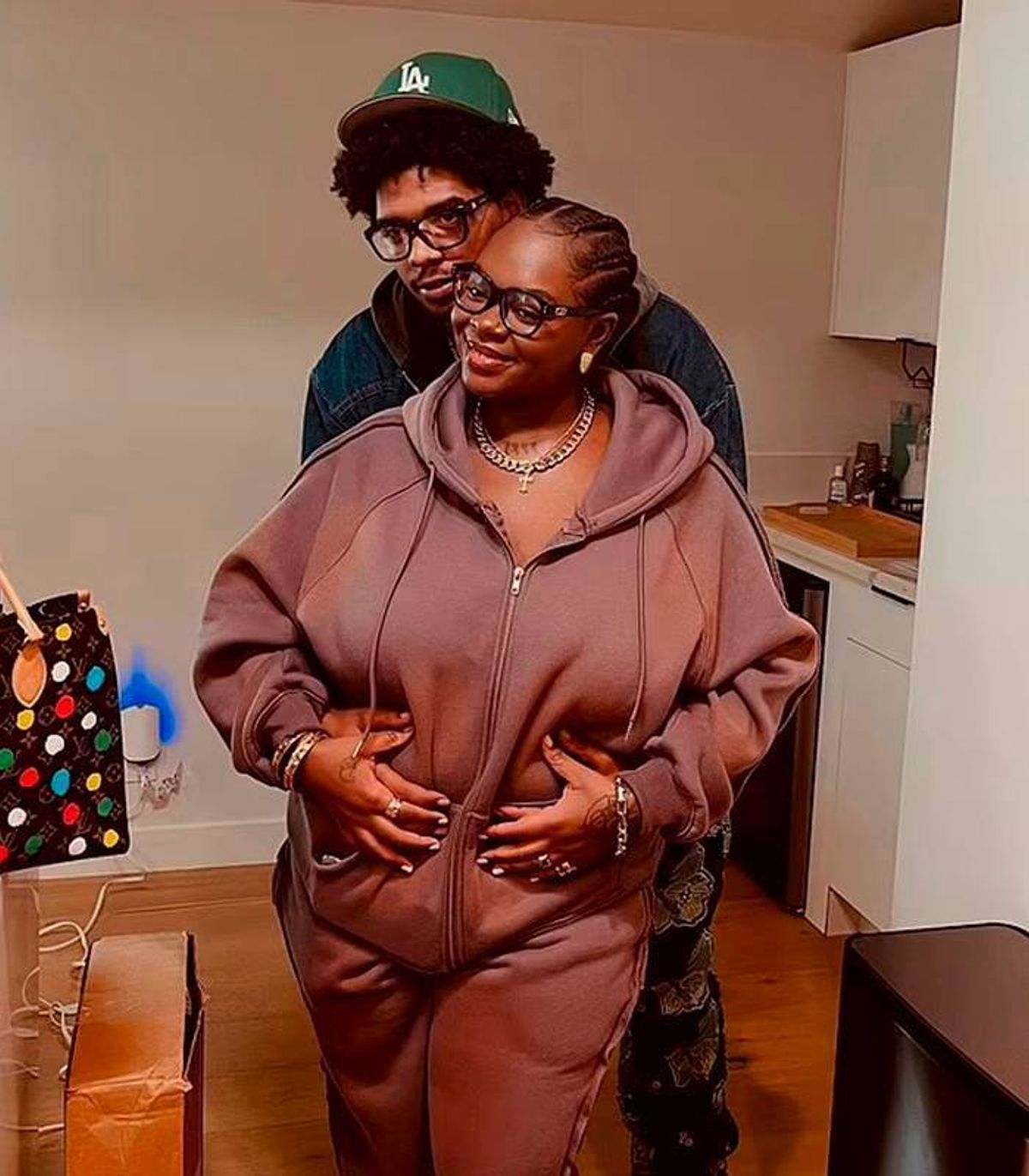 An individual stands in an indoor setting, dressed in a brown hooded garment, with their hands resting on their abdomen. Another person, partially visible in the background, stands behind them. The scene takes place in a room with white walls and a wooden floor, with various household items in the background.