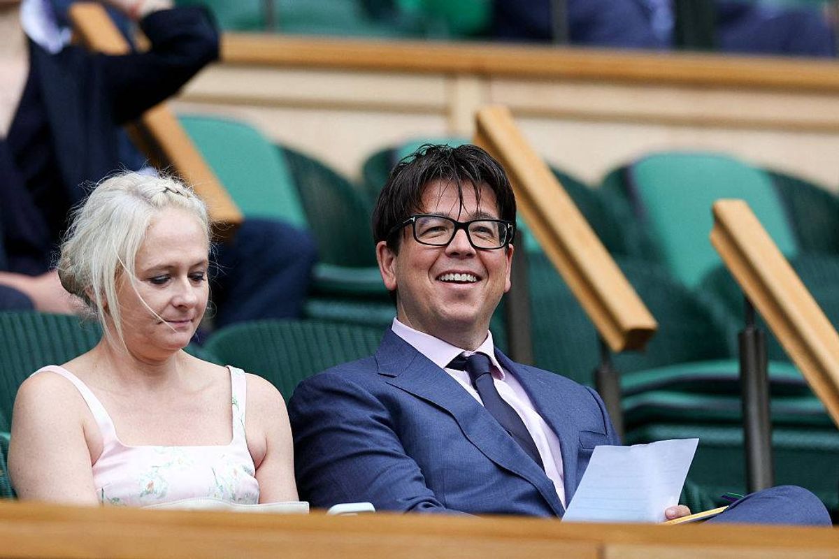 An individual in formal attire, seated in a green stadium seat, is accompanied by another individual. They both appear to be smiling and engaged in conversation.