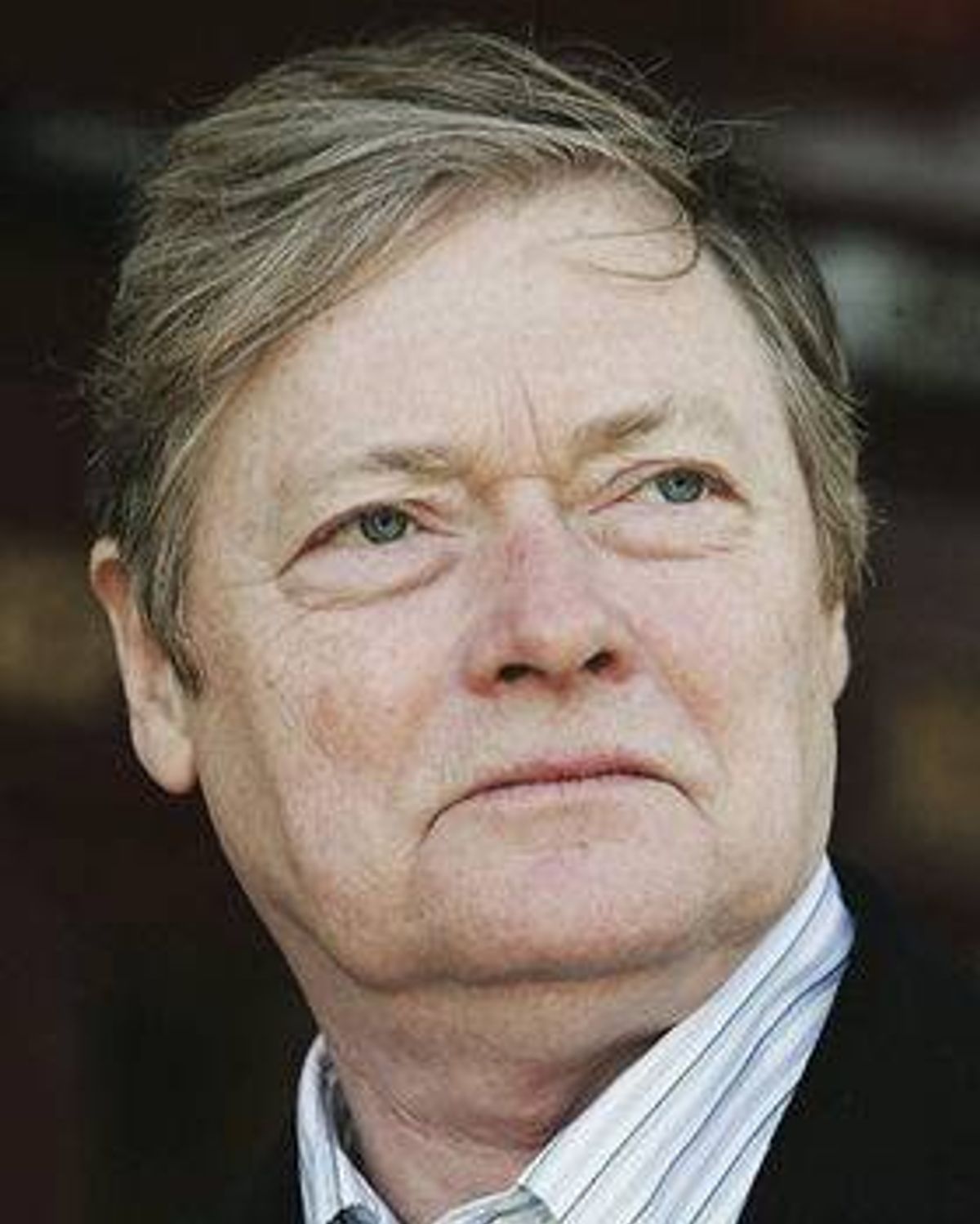 A close-up portrait of a distinguished individual with a serious expression, dressed formally in a collared shirt and dark blazer, set against a softly blurred background.