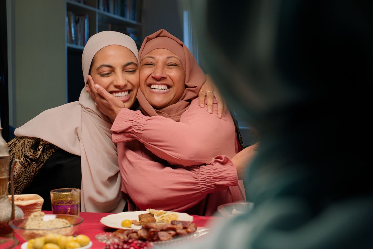 Women, hug and food for Ramadan celebration, Muslim people at dinner table together eating, laughing and smile. Islam, embrace and family with culture, feast or happy festival of eid al fitr in home
