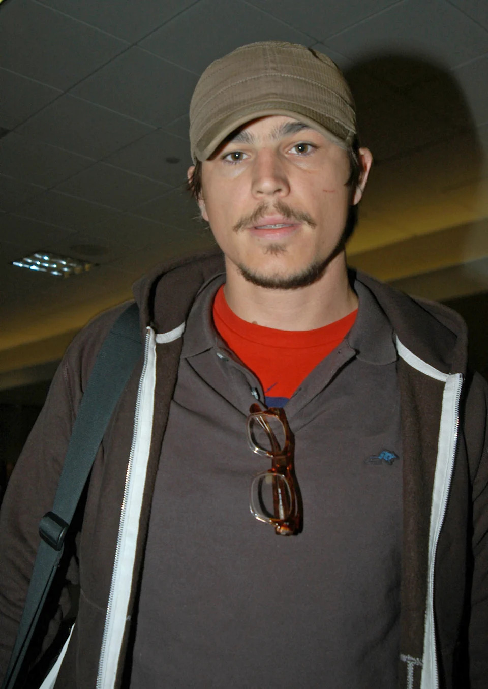 A person with a mustache and beard wears a cap, hoodie, and glasses hanging from their shirt inside a building