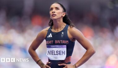 Laviai is stood with her hands on her hips looking up. She is wearing a GB navy tight fitting running vest. The words Great Britain are printed in red on her chest. She has a name tag with Nielsen on her front. She has dark brown curly hair tied off her face.