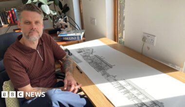 A man with grey and brown hair slicked away from his forehead is sat in a chair next to a desk looking at the camera. He has his hands together and knee crossed and is wearing a brown short-sleeved shirt and blue trousers. On the table is a long paper roll with detailed drawings of buildings.