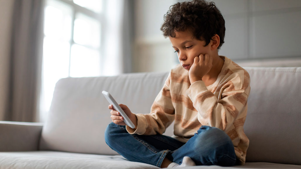 an elementary school student sits on a sofa looking at a smartphone