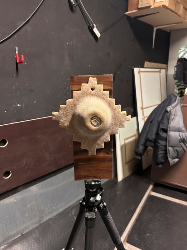 A geometric, textured sculpture resembling a stylized gear or abstract sun is mounted on a wooden board attached to a tripod. The background shows a coat, stacked boxes, and various objects in a cluttered room.