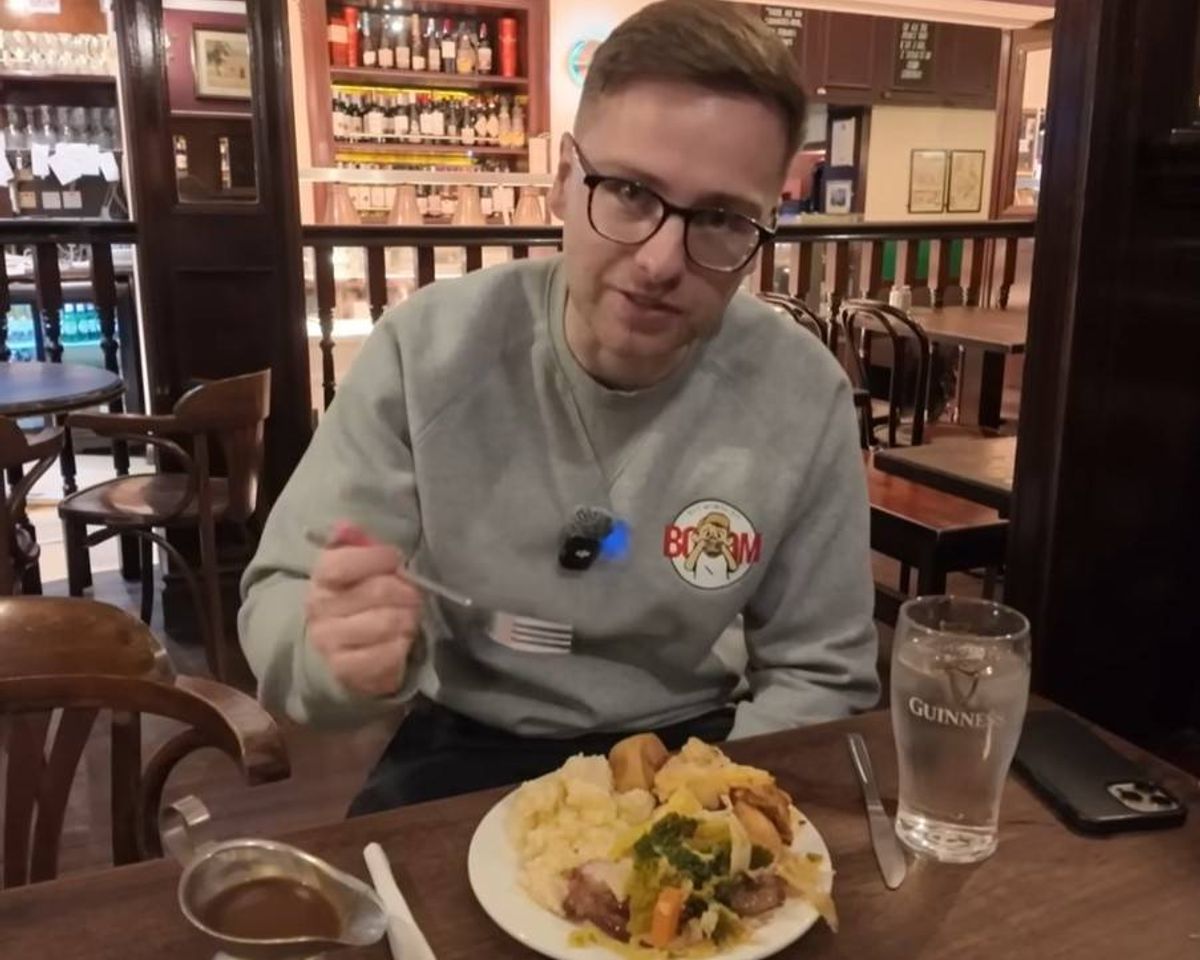 An individual wearing a gray shirt and glasses is seated at a table in a restaurant, holding a fork and spoon, and appears to be engaged in a meal. Various dining tables and chairs are visible in the background, along with a glass of liquid on the table.