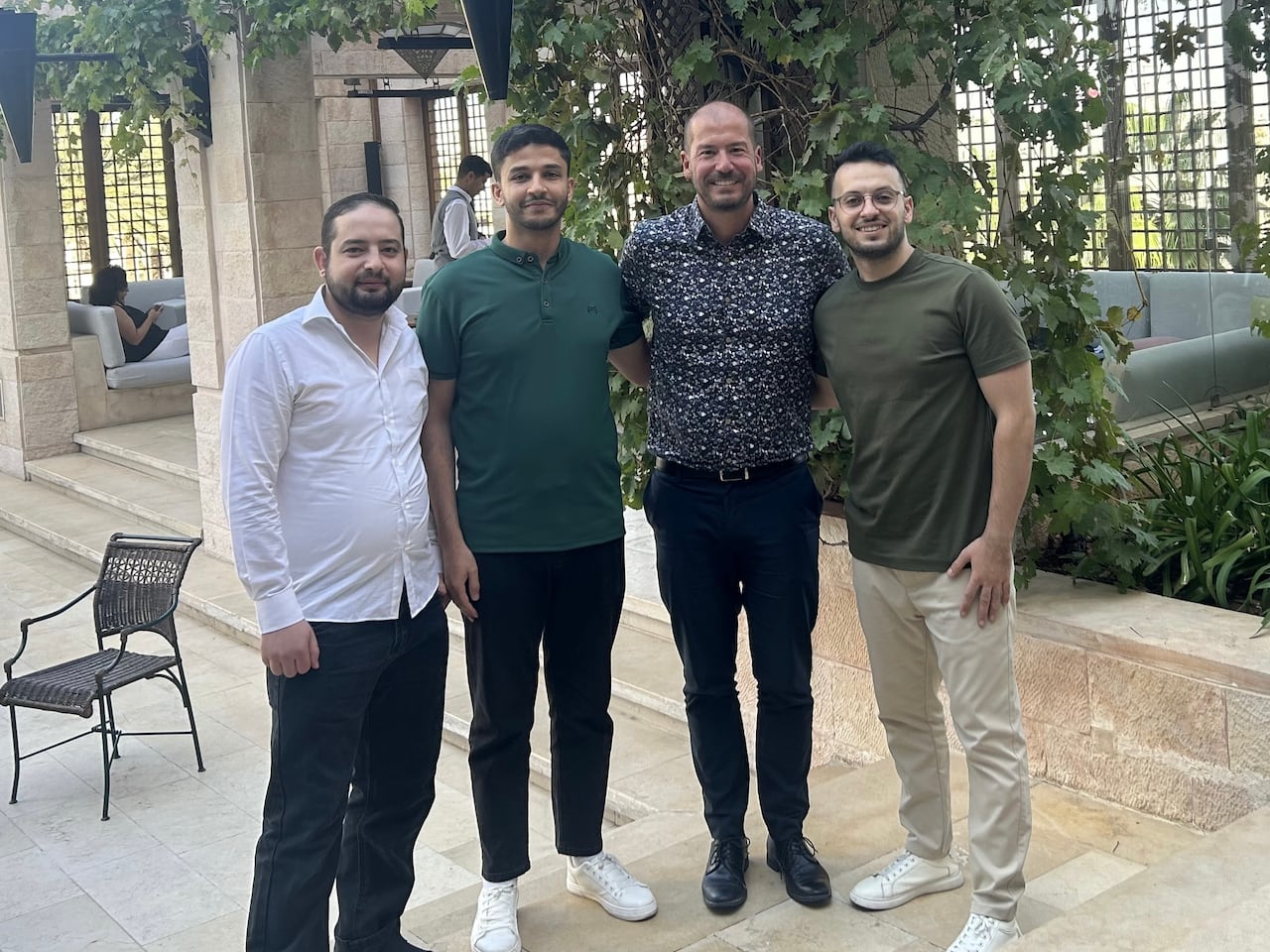 Four men are shown smiling in front of a white stone building with climbing plants.