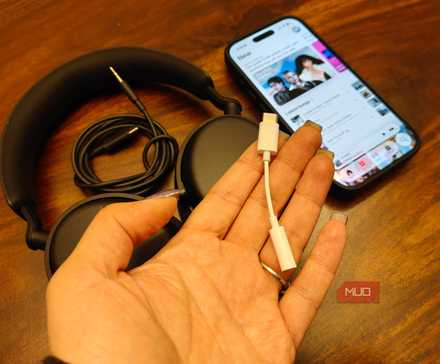 A hand holding a headphone adapter above a phone and headphones.