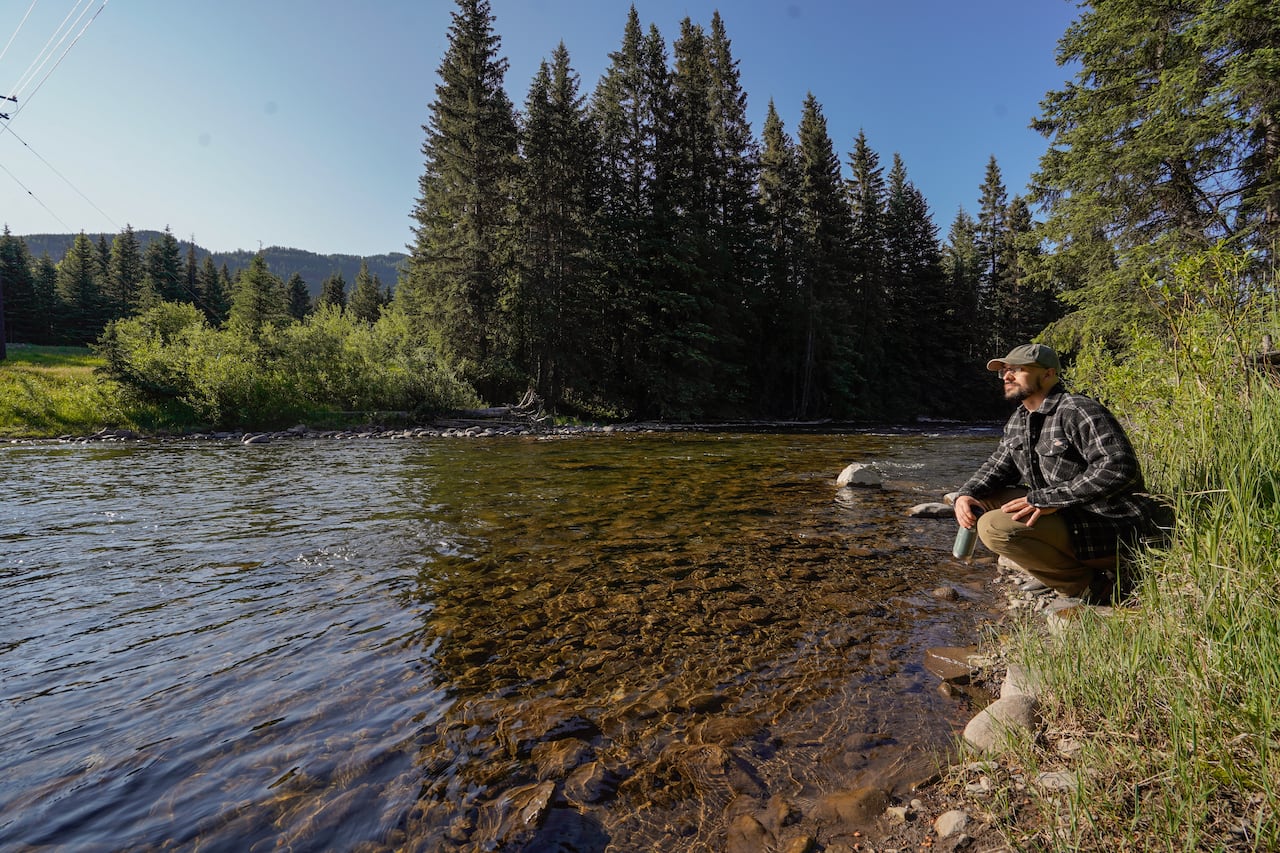 The image shows a man crouching at the edge of a shallow, clear mountain river in a forested valley.