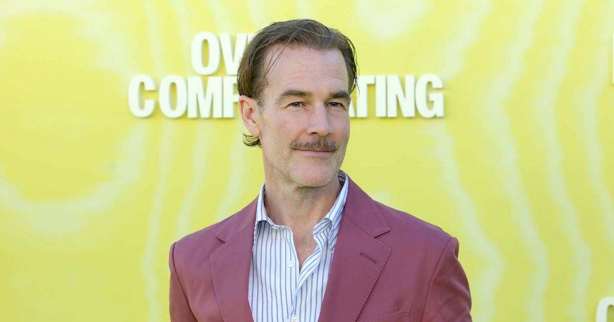 An individual dressed in a burgundy suit and light blue shirt stands against a bright yellow backdrop, likely at a public event or premiere. The background features the text "COMING" and "COM" in white font.