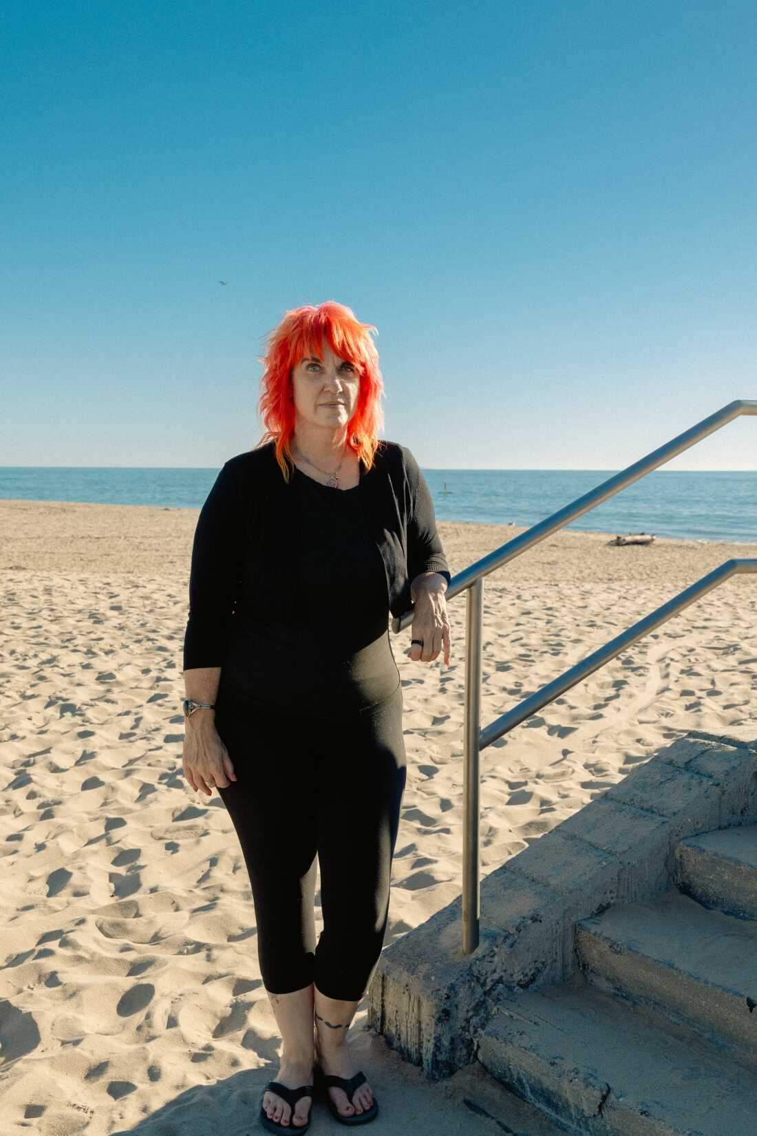 Micky Small stands on the beach at the base of cement stairs with sand and the ocean behind her.
